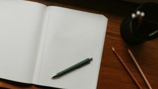 明るい木の机、ドライフラワーとマグ。ノートに書いている手