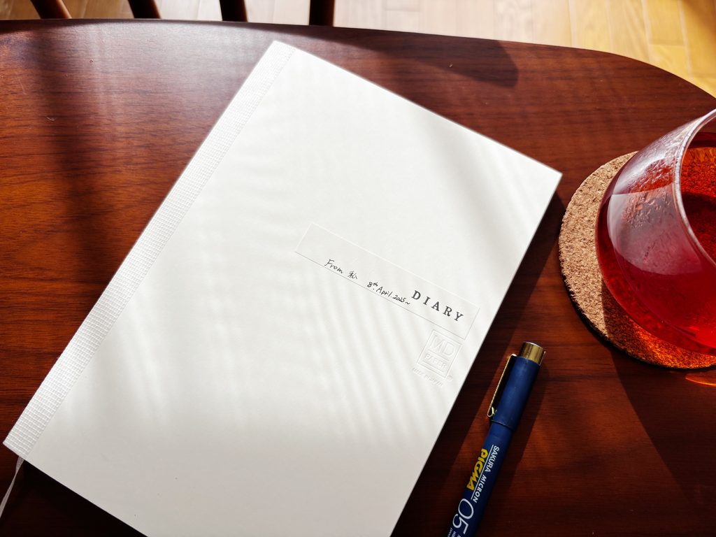 窓辺の光が差し込む木のテーブルに置いた、日記を書いているMDノート(A5・方眼)とペン、紅茶のグラス