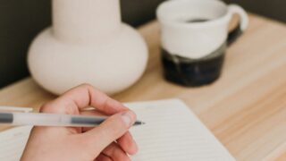 明るい木の机、ドライフラワーとカップ、ノートに書く手